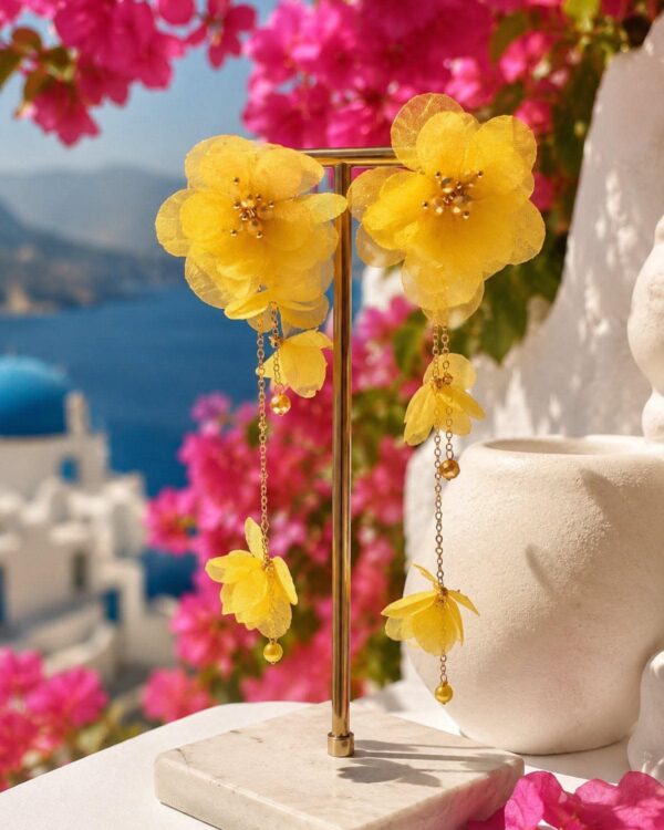 Yellow fabric-flower earrings on a gold display stand, with pink bougainvillea and blue sea background in a bright outdoor setting.