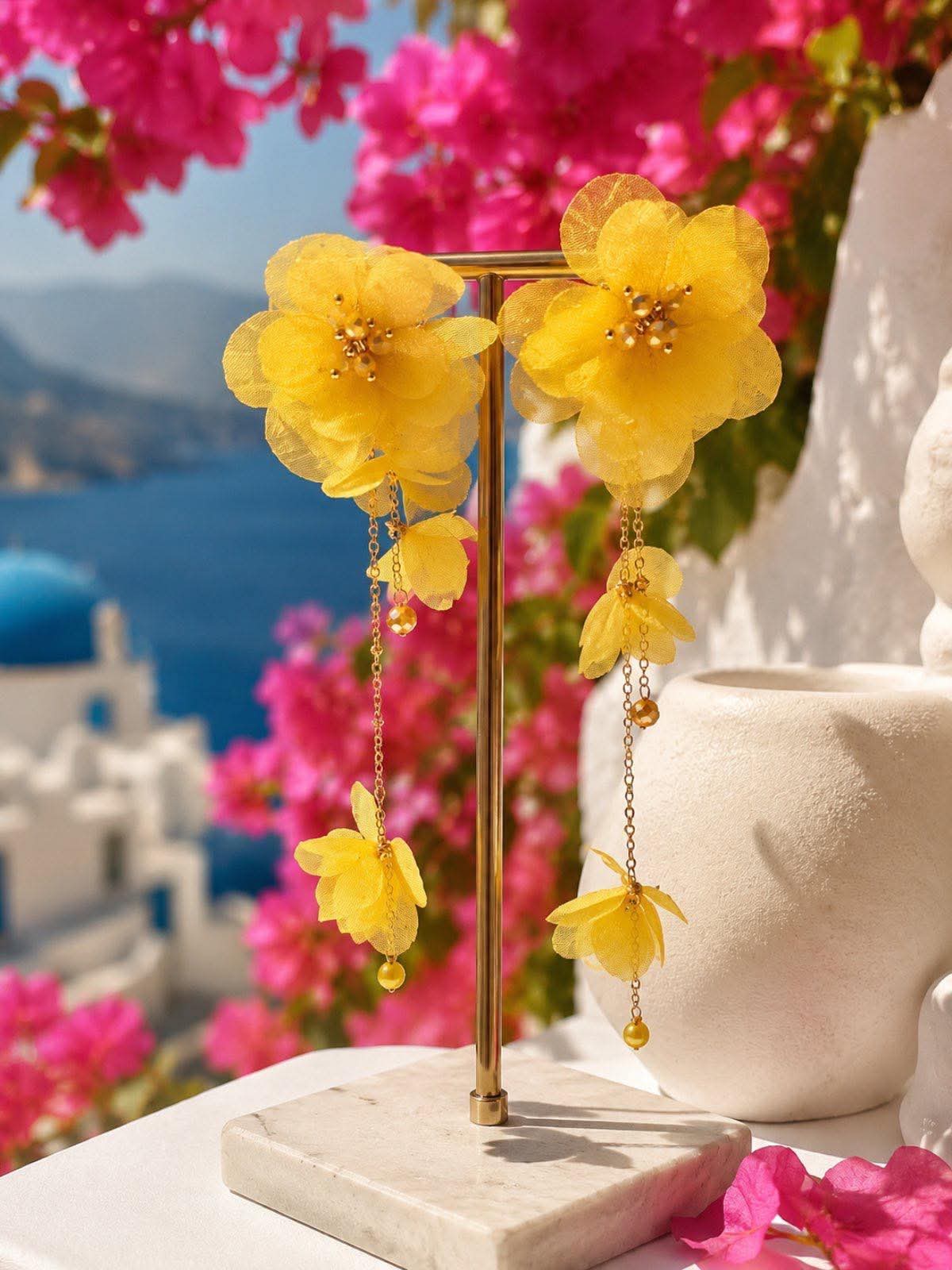 ηυτθυ Yellow fabric-flower earrings on a gold display stand, with pink bougainvillea and blue sea background in a bright outdoor setting.
