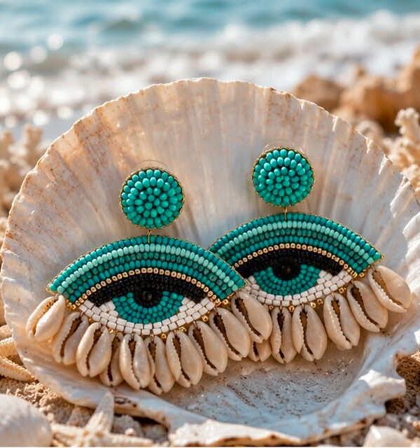 Turquoise beaded earrings shaped like eyes resting in a seashell on a sunny beach.
