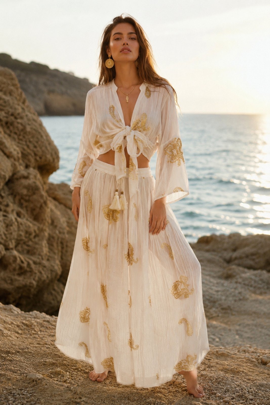 boho luxe set resort crop tom me desimo kai maxi fousta (1) Woman stands barefoot on rocky shoreline at sunset, wearing a white embroidered tie-front dress and gold jewelry by the sea.