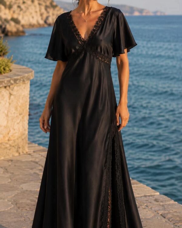 Woman in a long black satin gown with lace trim standing on a stone jetty by the sea at sunset, looking off to the side.
