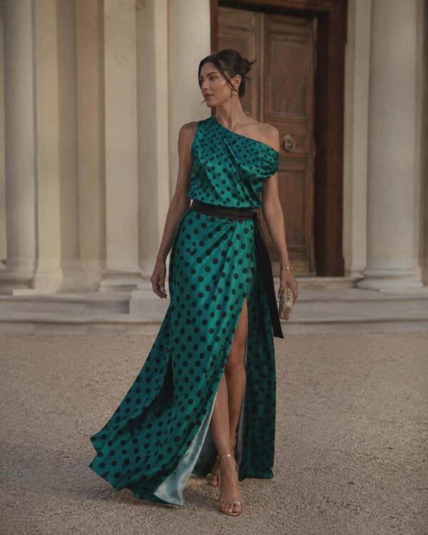Woman in a teal one-shoulder polka-dot gown with a high slit, standing before a neoclassical entrance of columns and a wooden door.