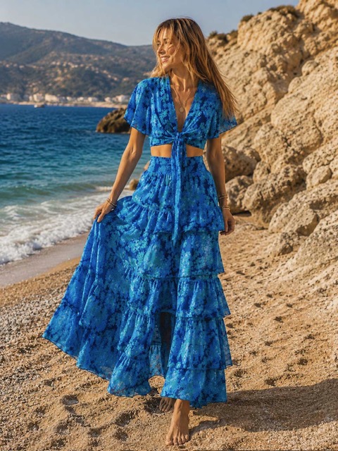 Woman walking barefoot on a rocky beach in a blue tiered maxi dress with a cutout at the waist.