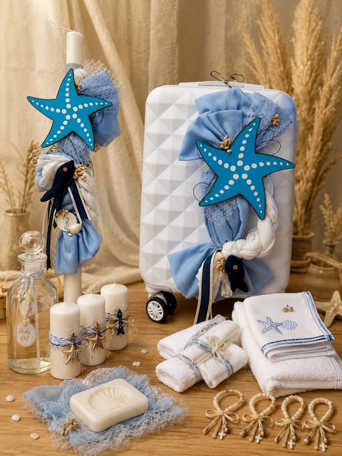 Coastal-themed display with a white geometric suitcase, blue ribbons and starfish ornaments, plus towels, soap, candles, and seashell decor on a wooden surface.