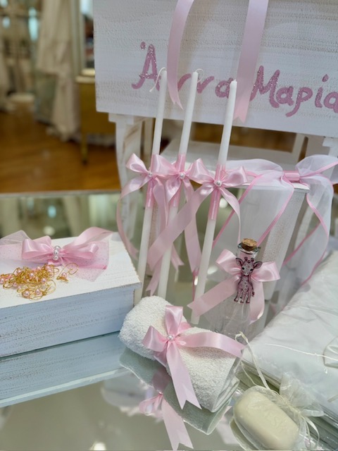 oloklhrwmeno paketo baptishs me ksulino mpoudouar me thema thn kamhlopardalh(6) Display of pink ribbon wedding decor: white boxes with pink bows, three white candles tied with pink ribbons, a small charm, and a fluffy white towel, in a shop display.
