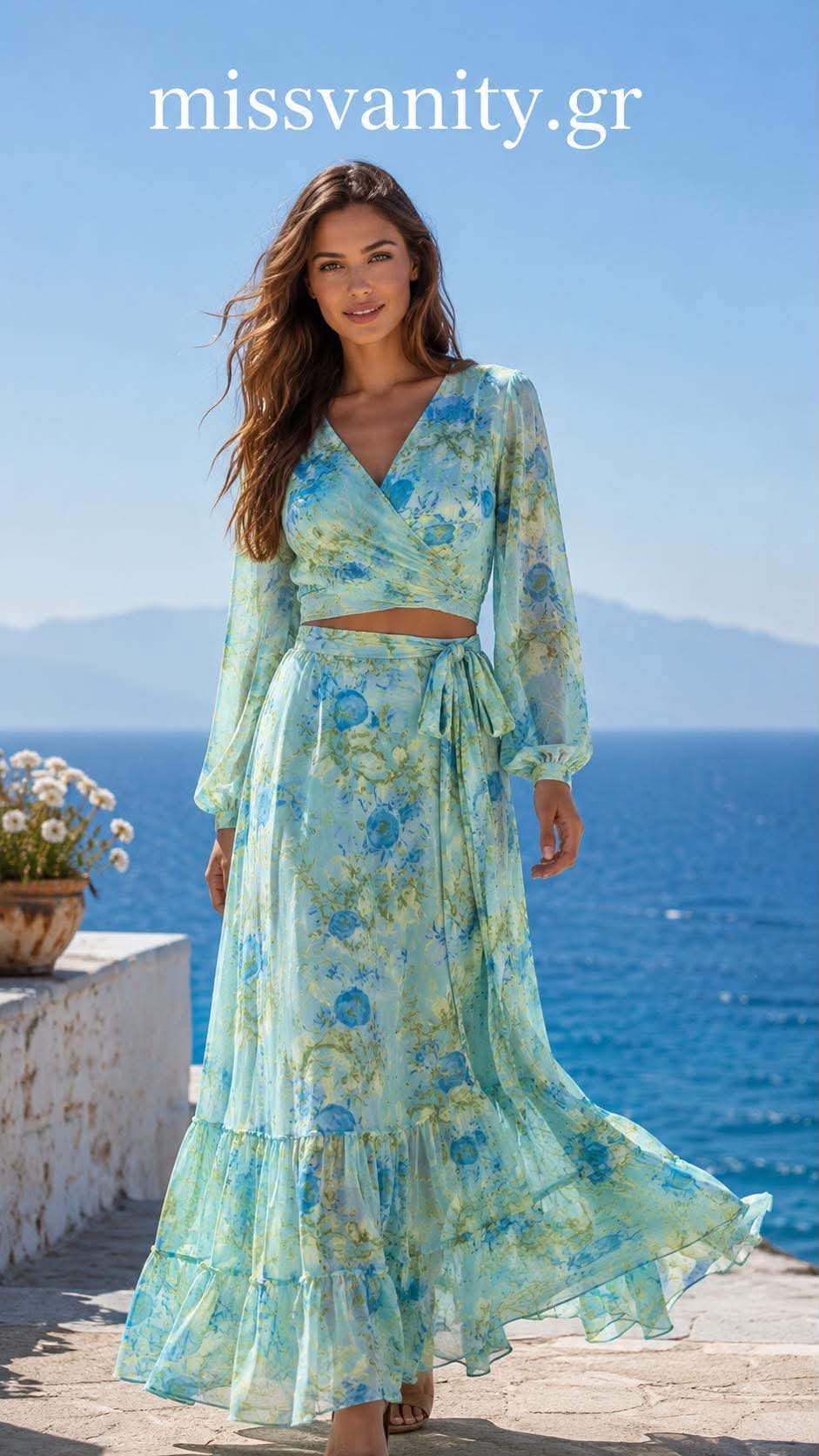 maxi floral set me krouaze crop top kai aerinh fousta(2) Model in a light blue floral maxi dress walking along a seaside walkway with the sea and mountains in the background, watermark 'missvanity.gr' visible at top.
