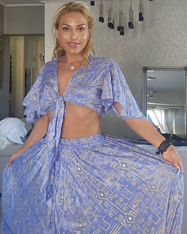 Woman with blonde hair in a blue geometric-print two-piece outfit, crop top tied at the front, standing in a bright, decorative room.