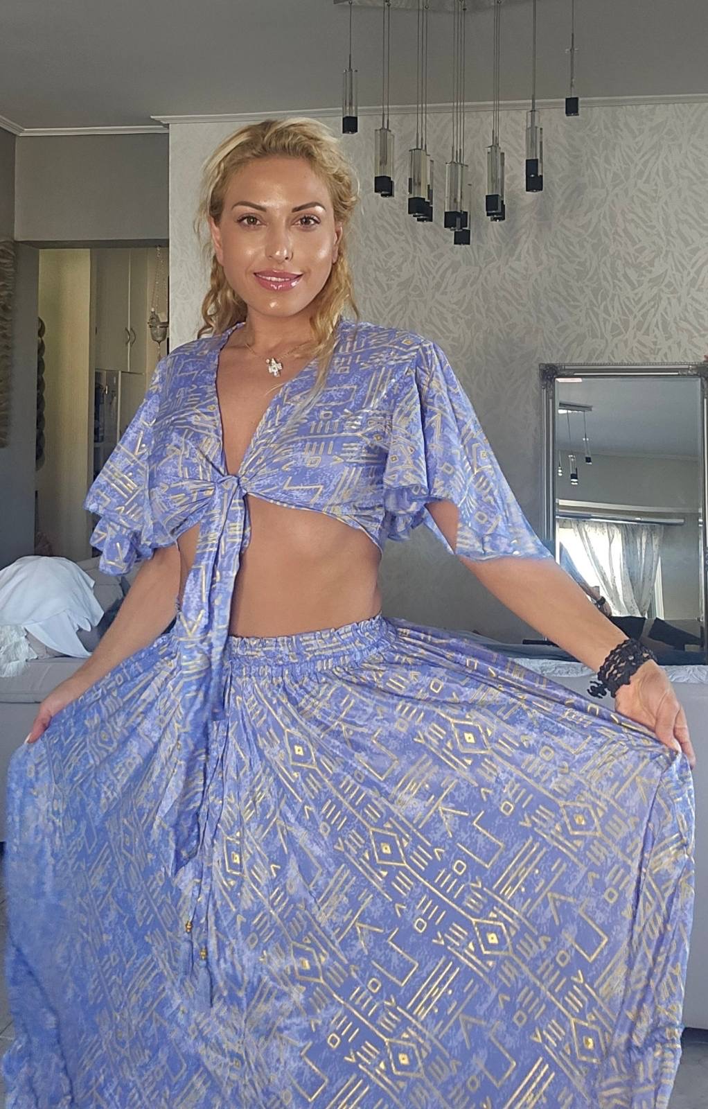 boho set me crop top kai fousta se polles apoxrwseis(1) Woman with blonde hair in a blue geometric-print two-piece outfit, crop top tied at the front, standing in a bright, decorative room.