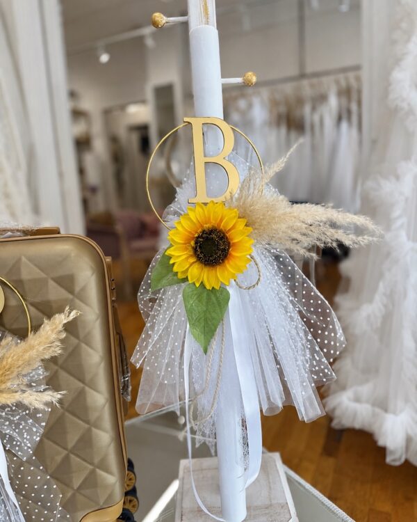 White candle centerpiece with a gold B monogram, bright sunflower, and tulle ribbons (wedding/party décor)