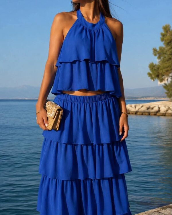 Woman by the water wearing a blue halter-tiered dress and sunglasses, holding a beige clutch.