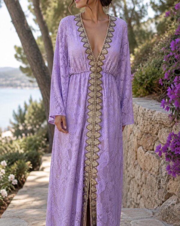 Woman in a lavender lace maxi dress with gold trim standing outdoors along a stone path, overlooking a coastal landscape.