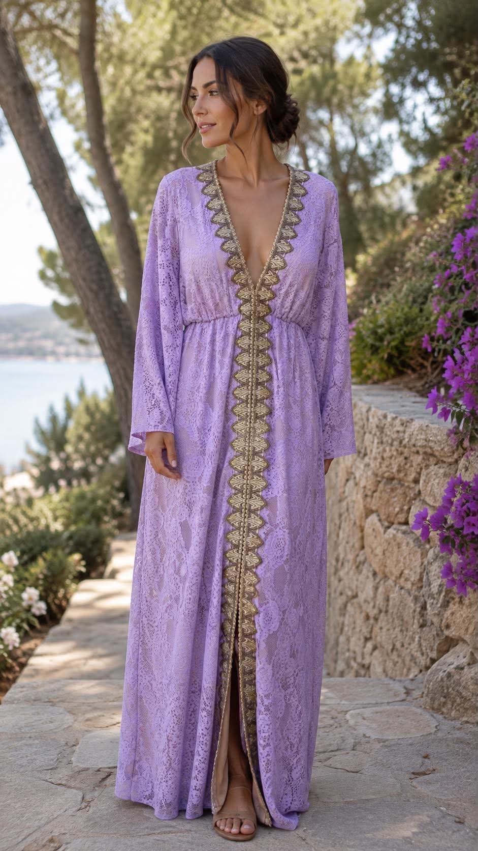 dantelenio maxi forema me xrysh tresa(1) Woman in a lavender lace maxi dress with gold trim standing outdoors along a stone path, overlooking a coastal landscape.
