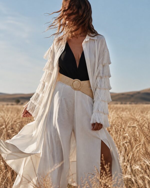 Woman in a white fringed coat over a black top and white wide-leg pants stands in a golden field with wind-blown hair.