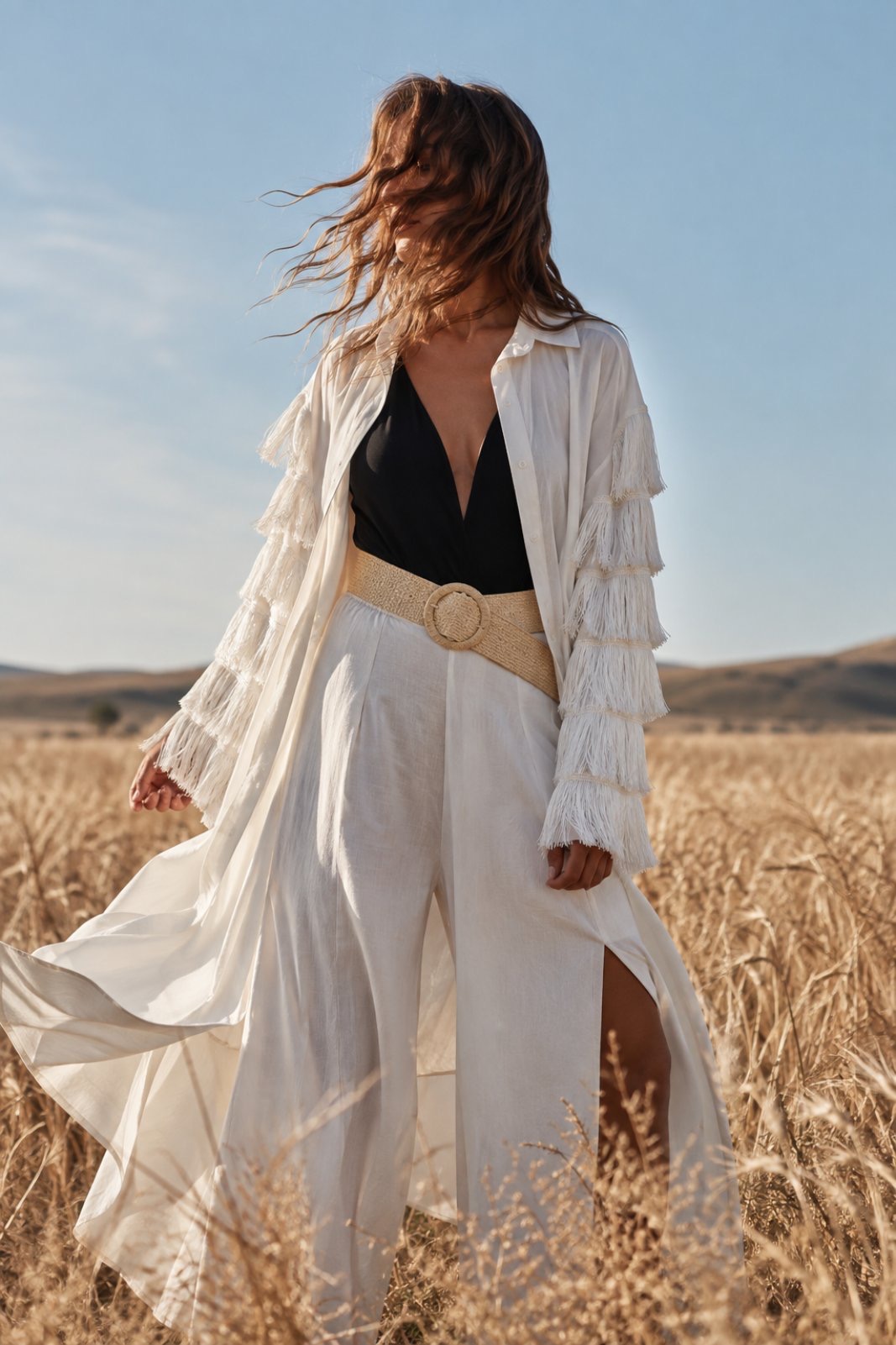 mouselina poukamiso me treses kai krosia(2) Woman in a white fringed coat over a black top and white wide-leg pants stands in a golden field with wind-blown hair.