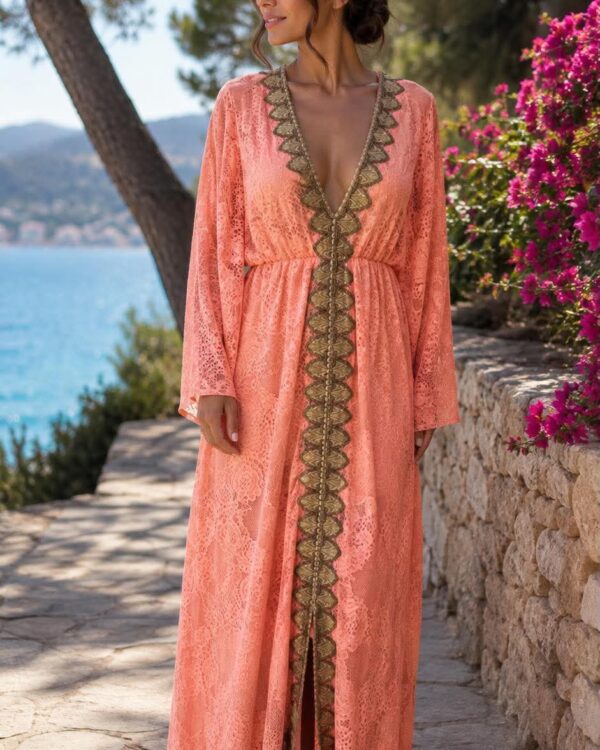 Woman in a peach lace maxi dress with gold trim, standing on a stone path by the sea, pink flowers nearby