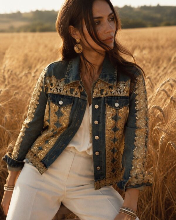 Woman posing in a golden-hour field wearing a decorated denim jacket with gold embroidery and large earrings, looking to the side.