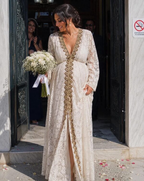 Bride in a cream lace gown with gold trim exits a doorway, holding a bouquet of white roses and baby's breath with a white ribbon.