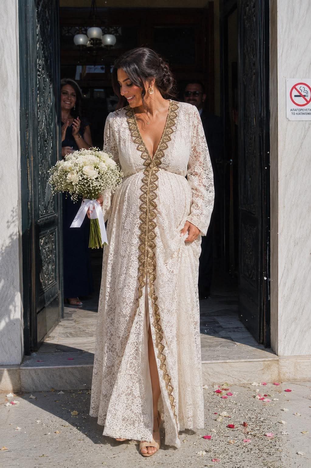 aerino nufiko forema dantela me xryses leptomereies(1) Bride in a cream lace gown with gold trim exits a doorway, holding a bouquet of white roses and baby's breath with a white ribbon.
