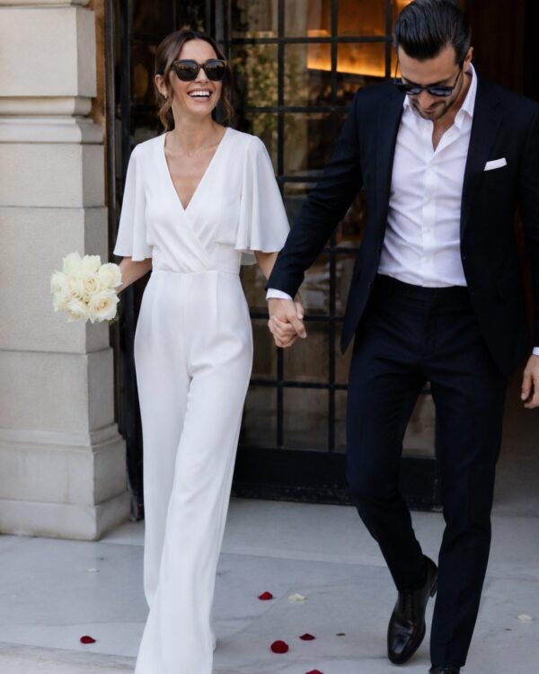 A smiling couple walking outside hand in hand; woman in a white jumpsuit holds a bouquet of white roses, man in a dark suit and sunglasses beside her.
