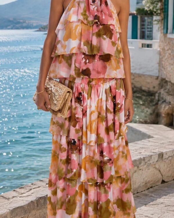 Woman in a layered pink, yellow, and green floral halter maxi dress by the sea, holding a woven clutch.